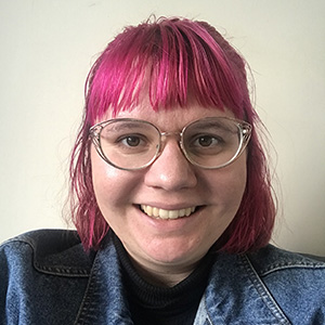 Maddy facing the camera, with pink hair in a bob, wearing rounded glasses and a denim jacket