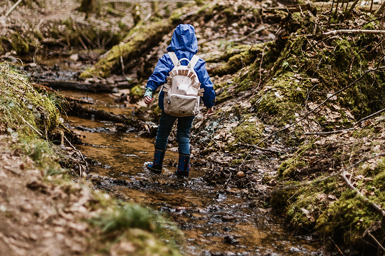 child wearing a rucksack shown from the back walking in wellington boots in a stream in the woods