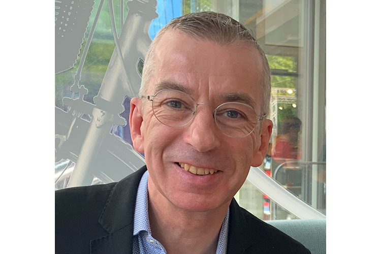 Paul Hibbert smiles at the camera, in a headshot taken against a background of windows and reflections. He is wearing a dark suit jacket and blue shirt.