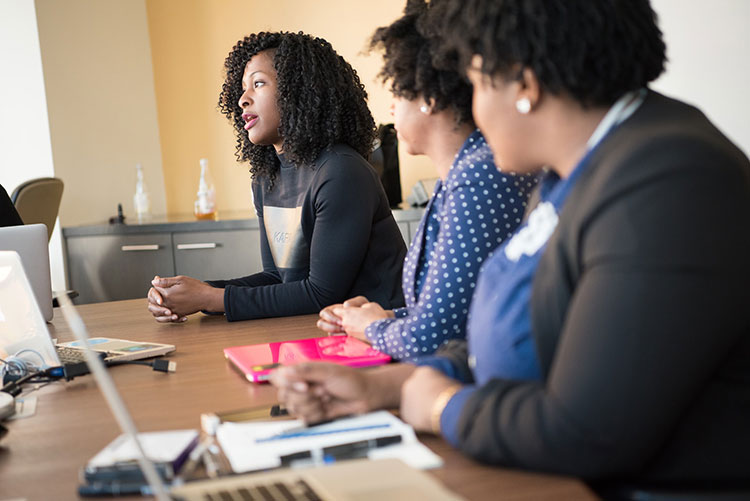 two women with laptops and notebooks sitting at table looking at woman talking