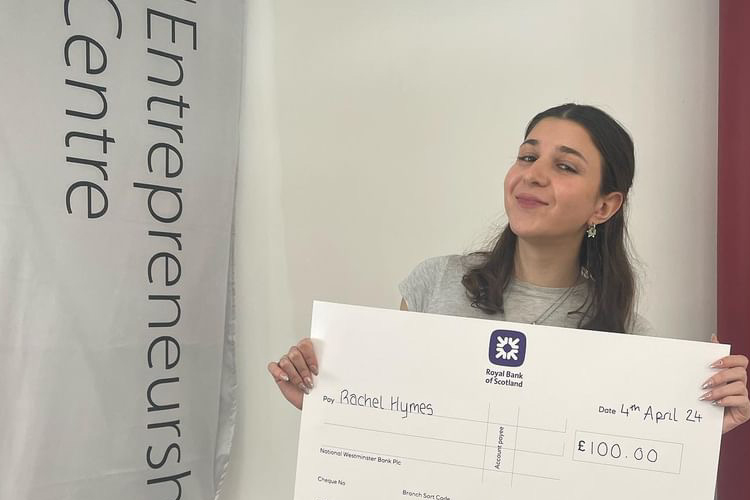 Rachel Hymes, smiling broadly and holding a big cheque for £100 made out to her, next to a banner with text 'Entrepreneurship Centre' partly visible