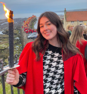 Hayley Stone, School President outside holding a lit torch, as part of the torchlight procession. Hayley is pictured in her red gown and other students can be seen walking along the historic pier behind her.